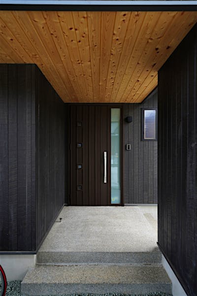 深い軒を設けた玄関ポーチ。黒い外壁は木目調サイディングで、木の風合いとメンテナンス性を兼ね備えている。天井の板張りとのコントラストも美しい。